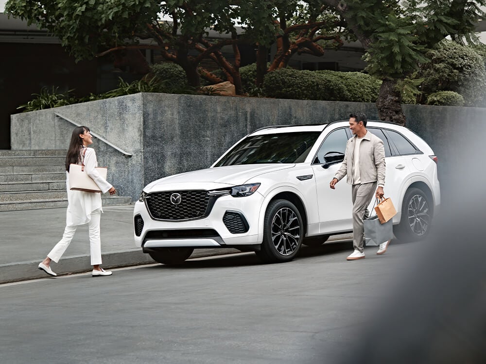 Two people walk beside a white CX-70 parked curbside.