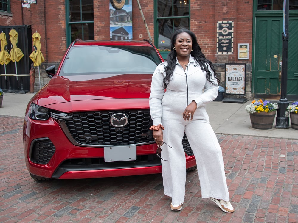 A woman in white stands in front of a red Mazda outside of a coffee shop.