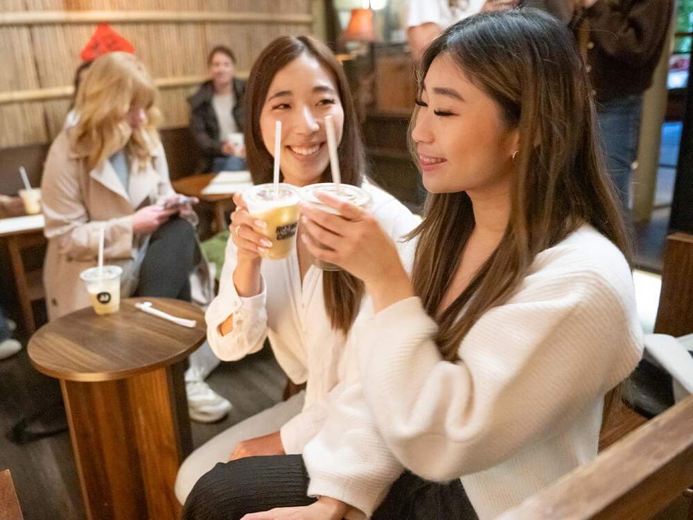 Two women hold up cups of coffee and smile while more people sit in the background.