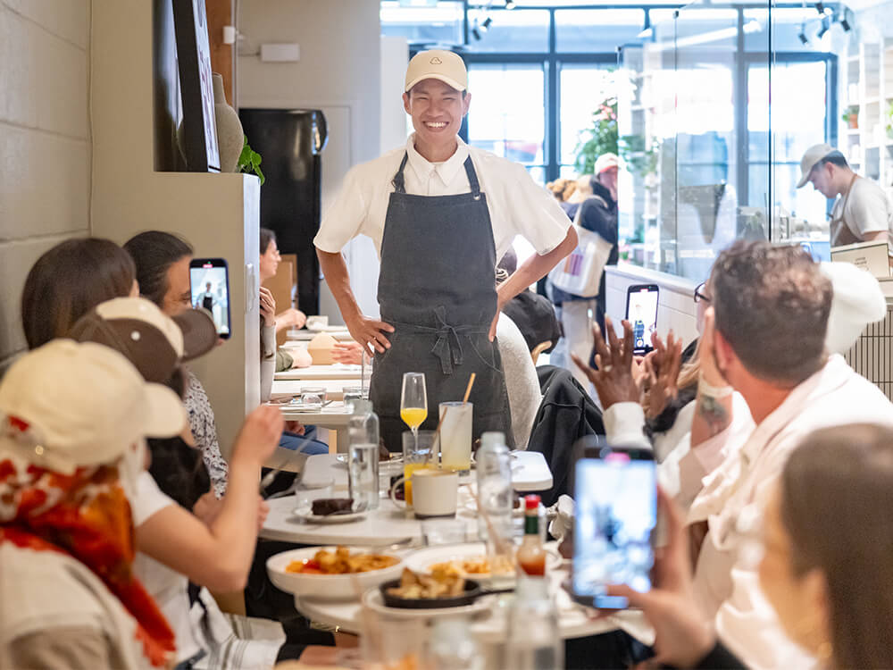 A smiling coffee shop employee stands in front of a table full of people eating and drinking.