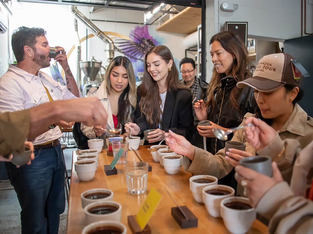 A group of people crowd around a table sampling several cups of coffee.