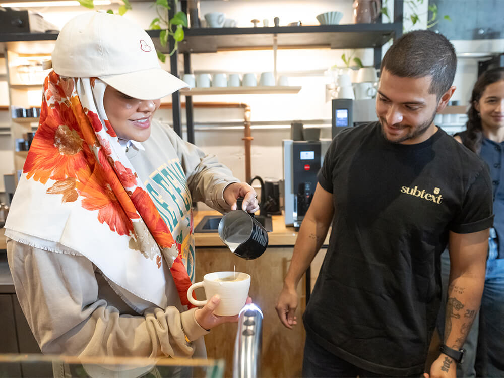 A smiling woman practices latte art in front of a coffee shop employee.