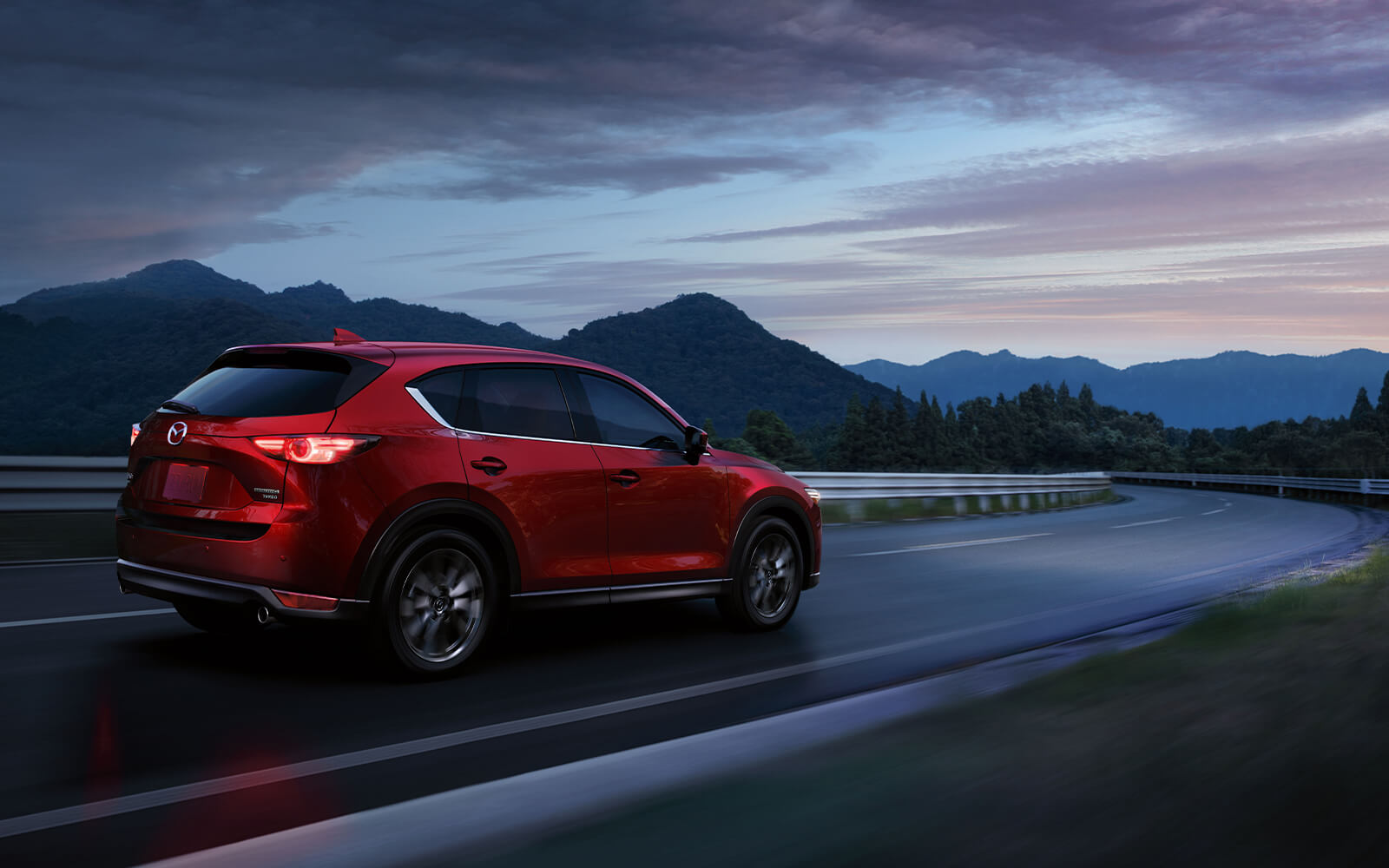 VUS Mazda rouge vibrant avec phares allumés dans une courbe sur une autoroute au crépuscule avec des montagnes sombres au loin. 