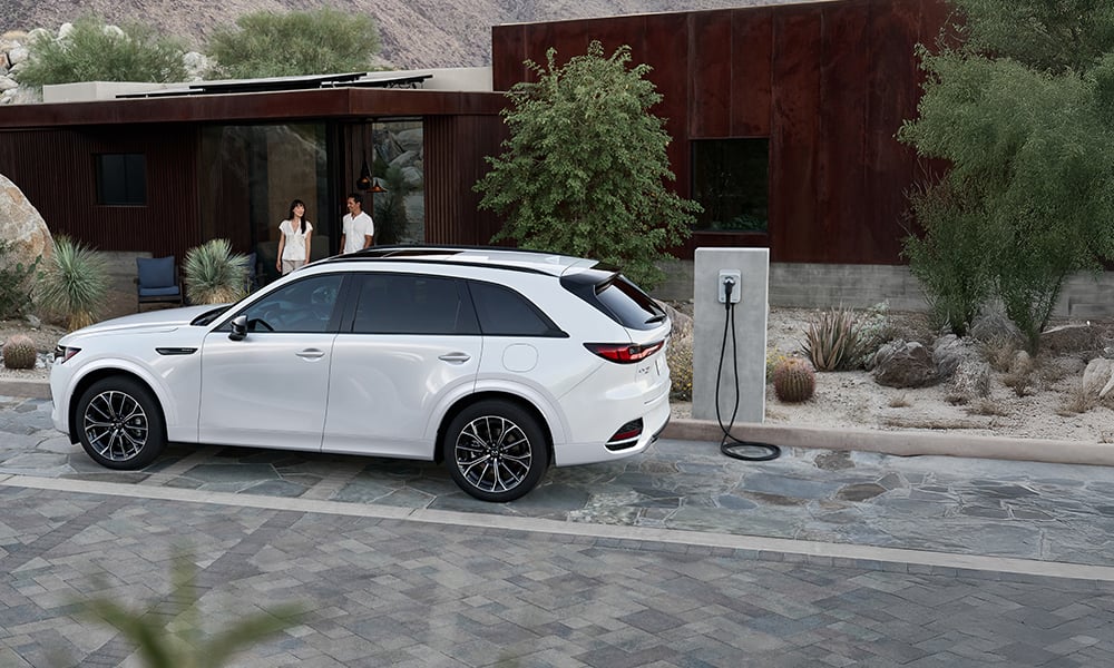 Silver Mazda MX-30 EV viewed from behind rear passenger side on beach with coastline on horizon.