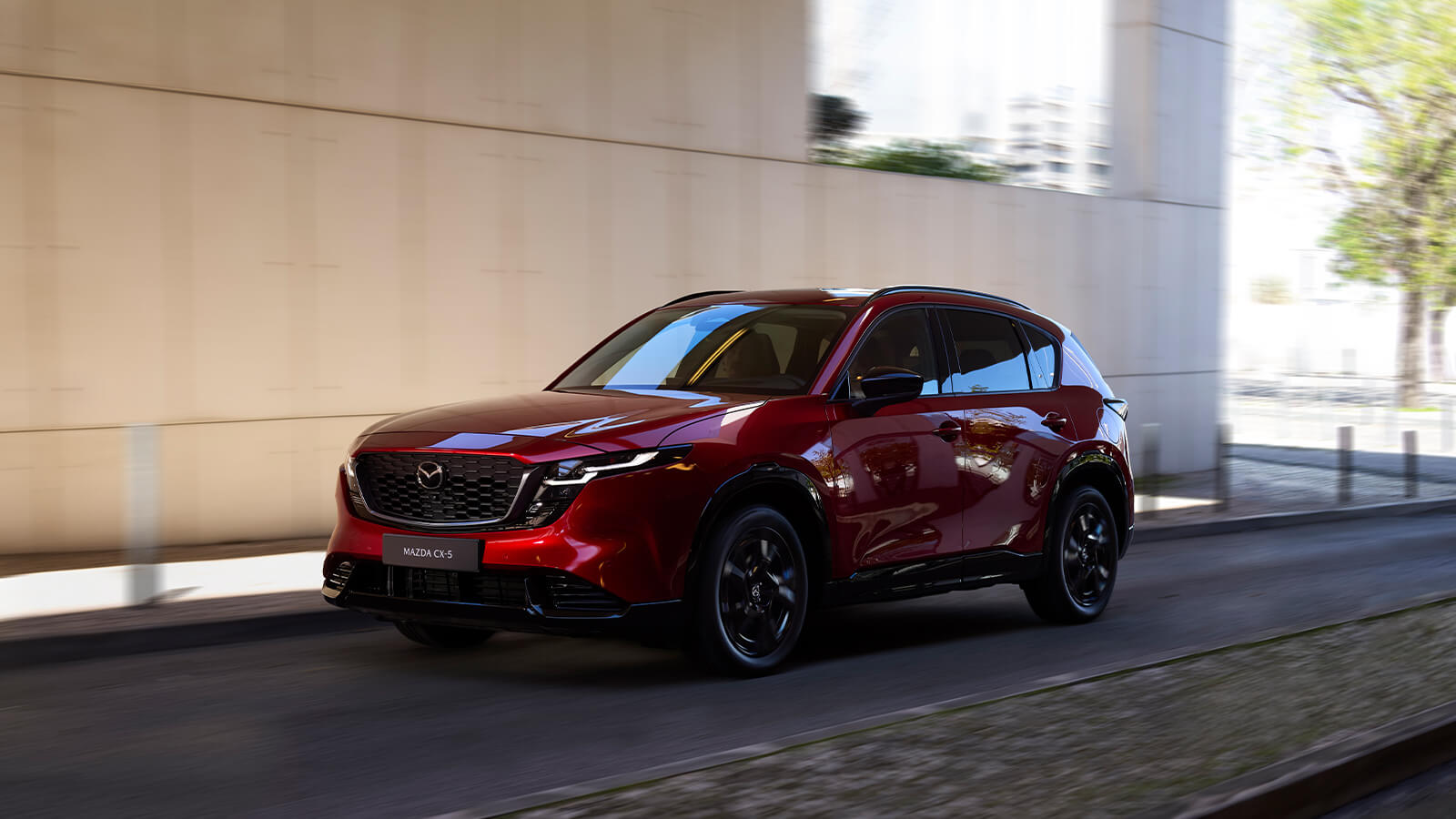 Profil latéral arrière d'un Mazda CX-5 rouge vibrant cristal, partiellement visible jusqu'à la roue arrière, garé dans une allée intérieure ombragée, avec en toile de fond une vue dégagée sur des montagnes lointaines.