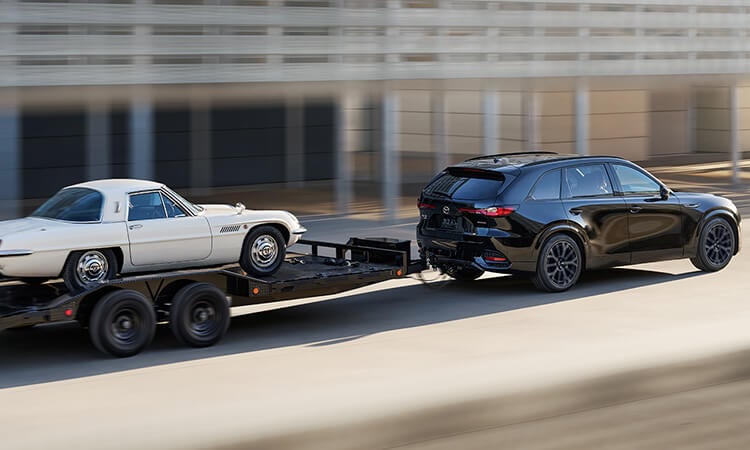 Jet Black Mica CX-70 towing a vehicle trailer hauling a white, classic Mazda leisure vehicle.