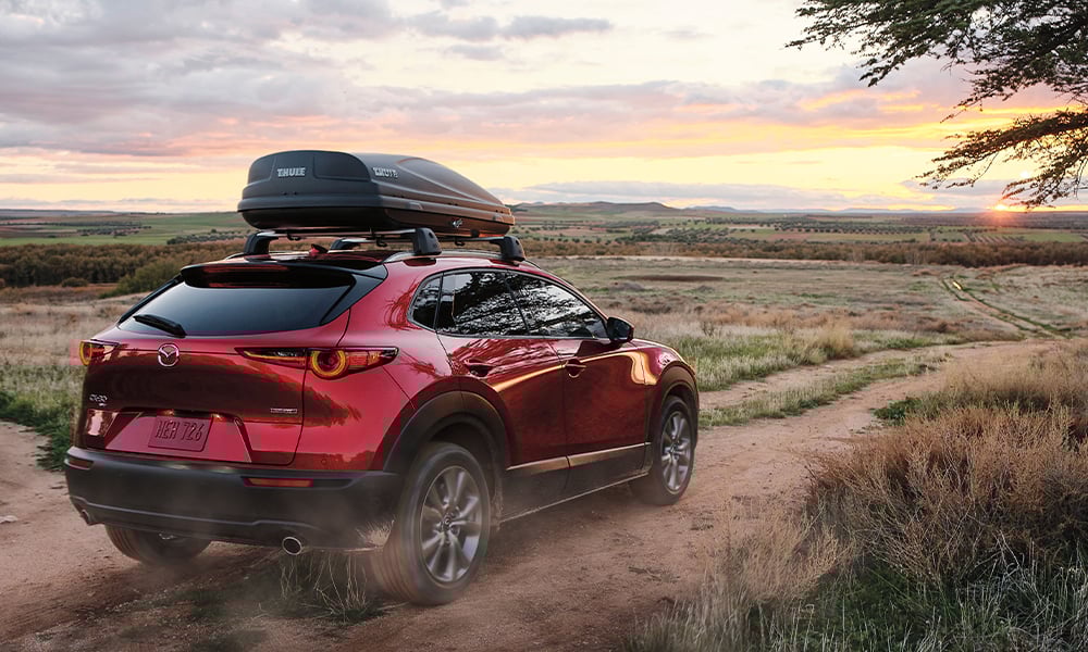 Man and woman stand near the trunk of a Soul Red Crystal Metallic Mazda CX-30 with a blurred outdoor landscape in the background.