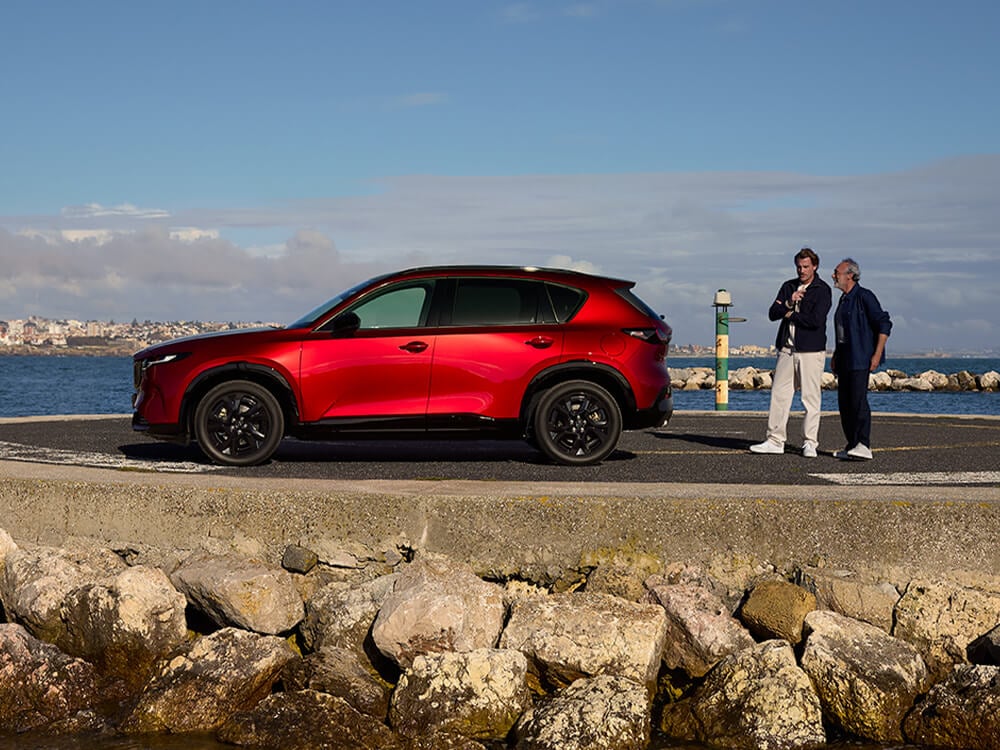 Driver side profile of a Soul Red Crystal Mazda CX-5 parked near a body of water, with two men standing near the rear of the vehicle.