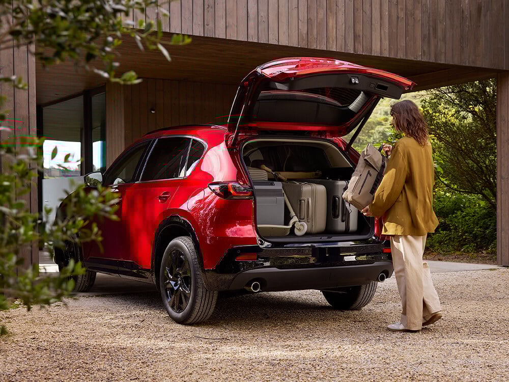 Rear three-quarter side profile of a Soul Red Crystal Mazda CX-5 in a driveway with the trunk open, as a woman places the last suitcase into a trunk already packed with luggage.