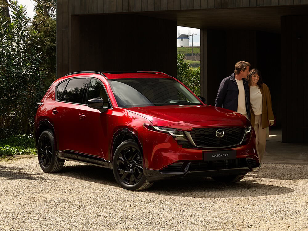 Front side profile of a Soul Red Crystal Mazda CX-5 parked in a driveway with a man and woman standing beside it.