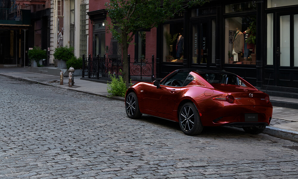 A red MX-5 RF with the top down sits parked on a city street next to storefronts.