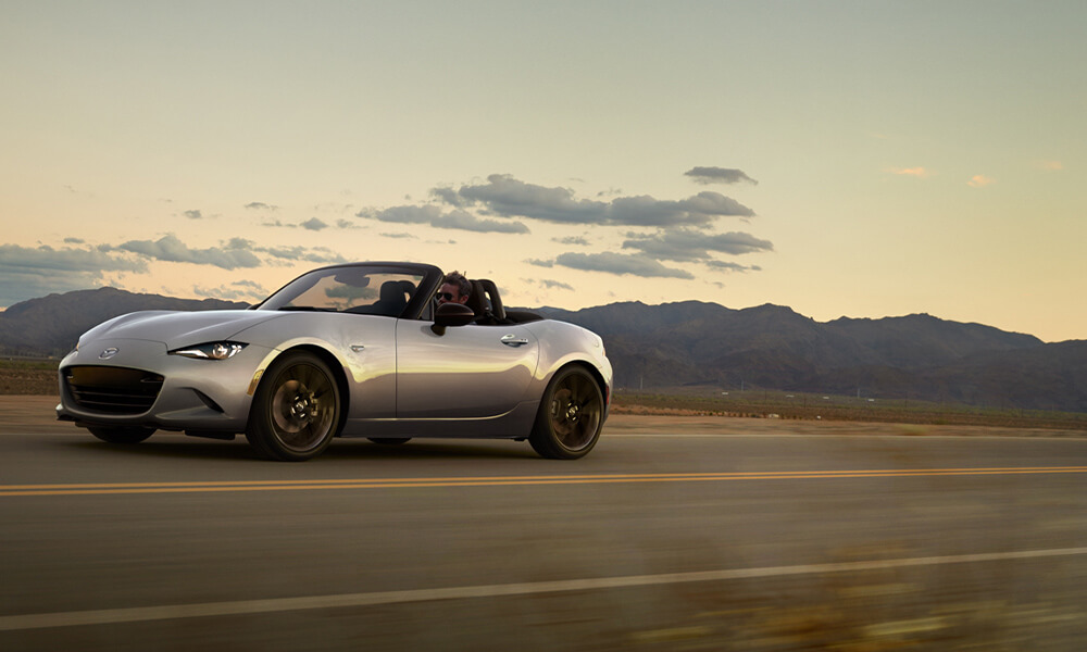 A silver MX-5 with the top down drives down an open road with mountains in the background.