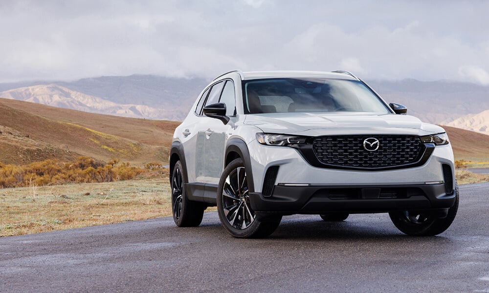 A white CX-50 Hybrid is on a road with cloudy skies and mountains in the background.