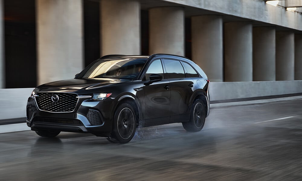 A black CX-70 Mild Hybrid Inline 6 Turbo drives down a street in front of a building with stone columns 