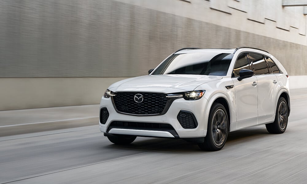 A white CX-70 Plug-In Hybrid drives down an industrial street next to a stone wall. 