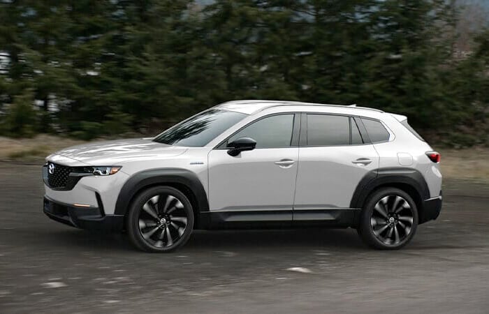 Un CX-50 hybride blanc roule sur une route bordée d’arbres. 