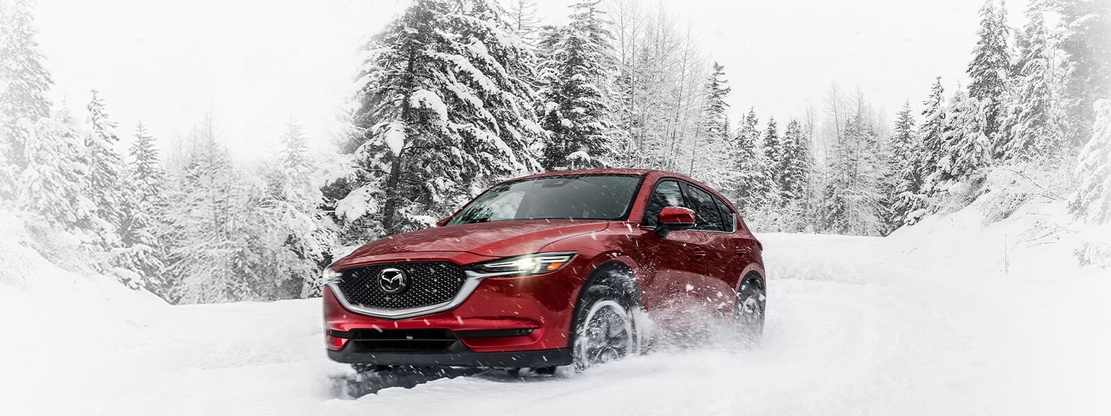 Un VUS Mazda rouge vibrant cristal avec les phares allumés passe devant des conifères enneigés alors qu’il négocie une courbe dans la neige profonde sur une route rurale 