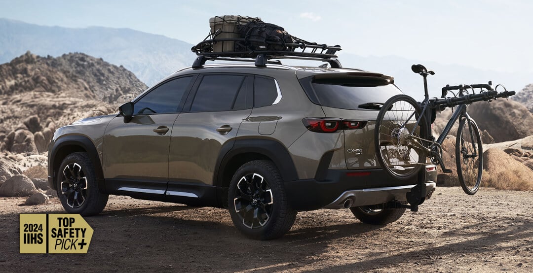 Rear view of Zircon Sand Metallic Mazda CX-50 parked, with roof rack loaded with outdoor gear, and bike rack holding a mountain bike. 