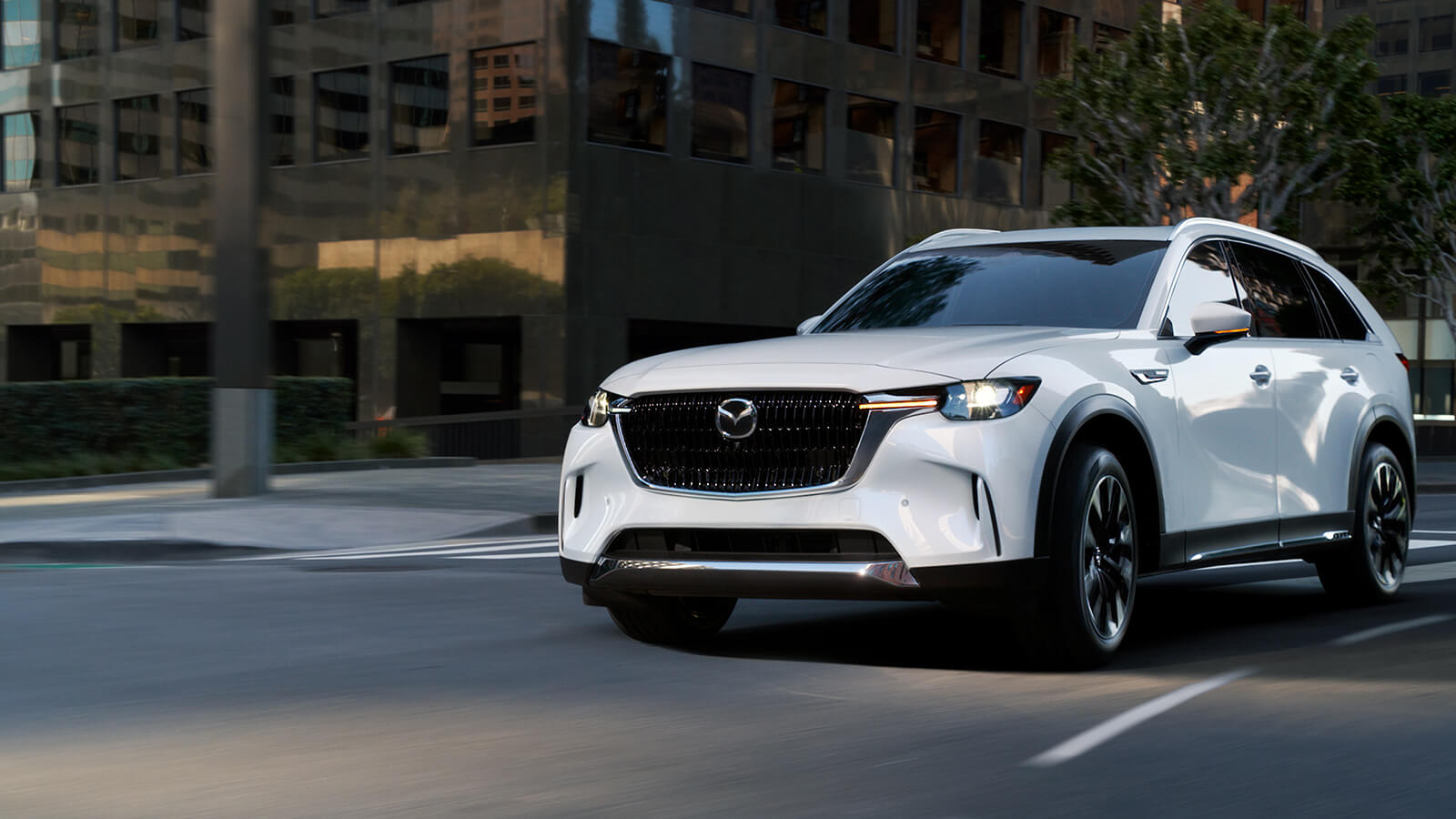 Une Mazda blanche devant un passage pour piétons dans une rue urbaine avec des bâtiments en arrière-plan. 