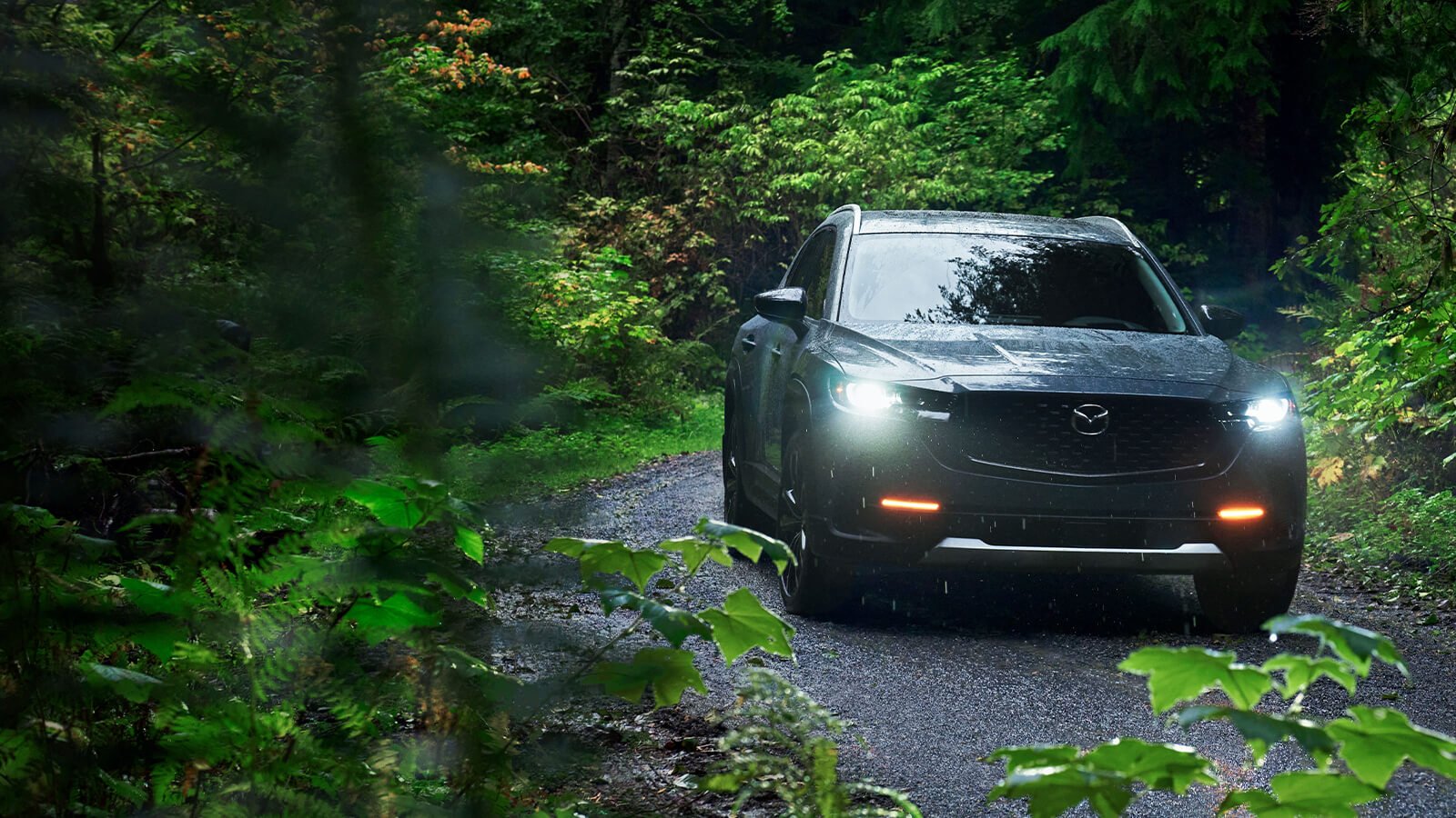 A Mazda with its headlights on drives down a road lined with green trees. 