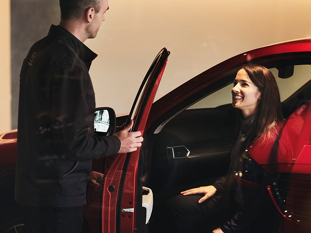 Un homme ouvre la portière d’une Mazda rouge. Une femme assise à l’intérieur de la voiture lui sourit. 