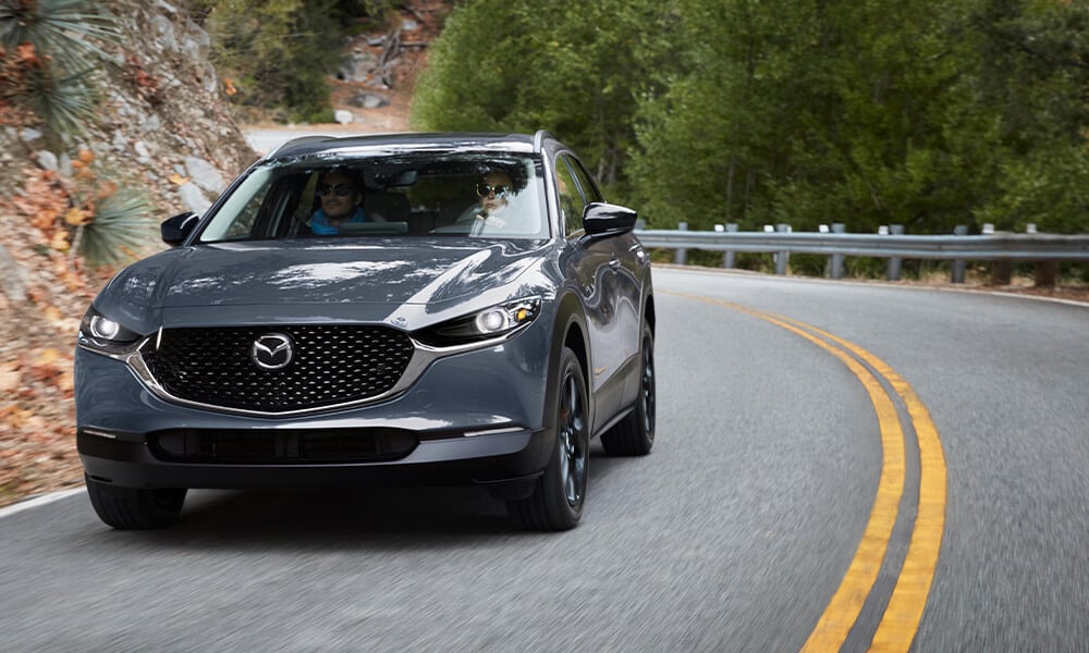 A dark grey Mazda drives along a curved road with trees on one side. 