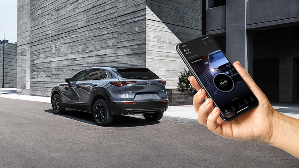 A hand holding a cellphone with the MyMazda app open, with a CX-30 shown in the background