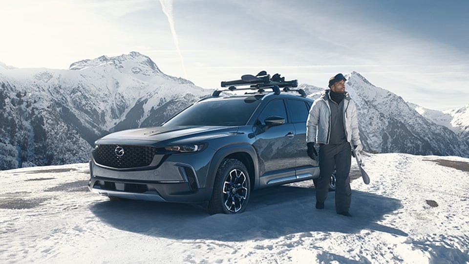 Man dressed for skiing holding a ski beside his Mazda CX-50 on top of a snowy mountain.