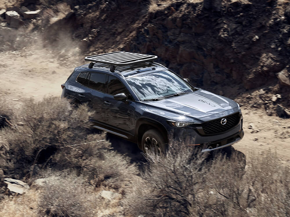 Overhead view of Mazda CX-50 with roof rack  trailing dust on dirt track between brush and rock.