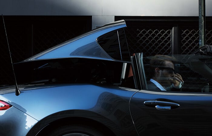 Un conducteur avec des lunettes de soleil qui baisse le toit de sa MX-5 RF gris mécanique métallisé garée.