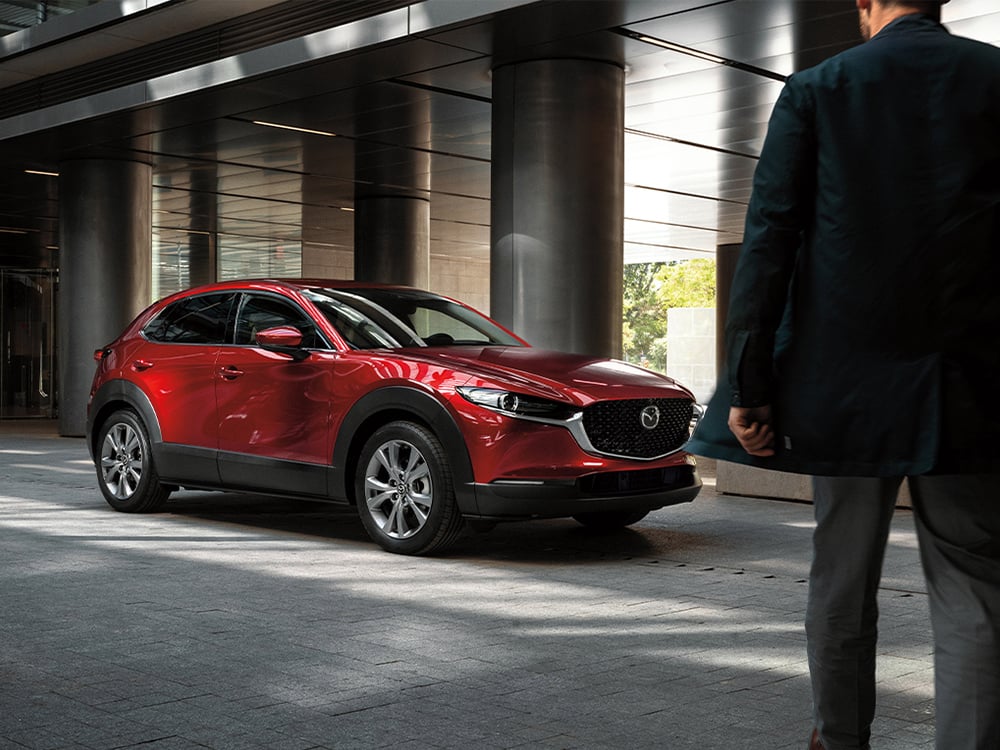 Man in foreground approaches Soul Red Crystal Metallic Mazda CX-30 parked ¾ front in drive of modernist building.