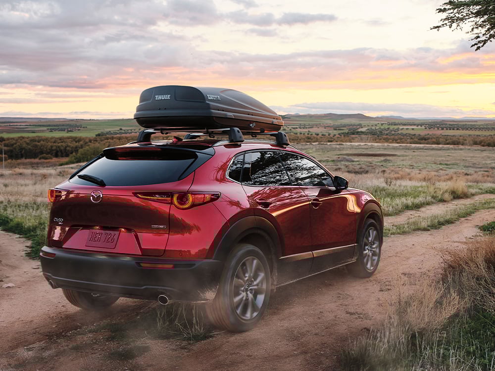 Back ¾ shot of Soul Red Crystal Metallic CX-30 with roof box, kicking up dust as it drives away on dirt path in rural-field environment.