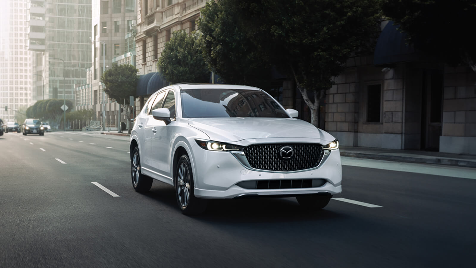 Close up of Rhodium White Metallic Mazda CX-5 ¾ front shot driving on wide city street, urban architecture and stately buildings in background. 