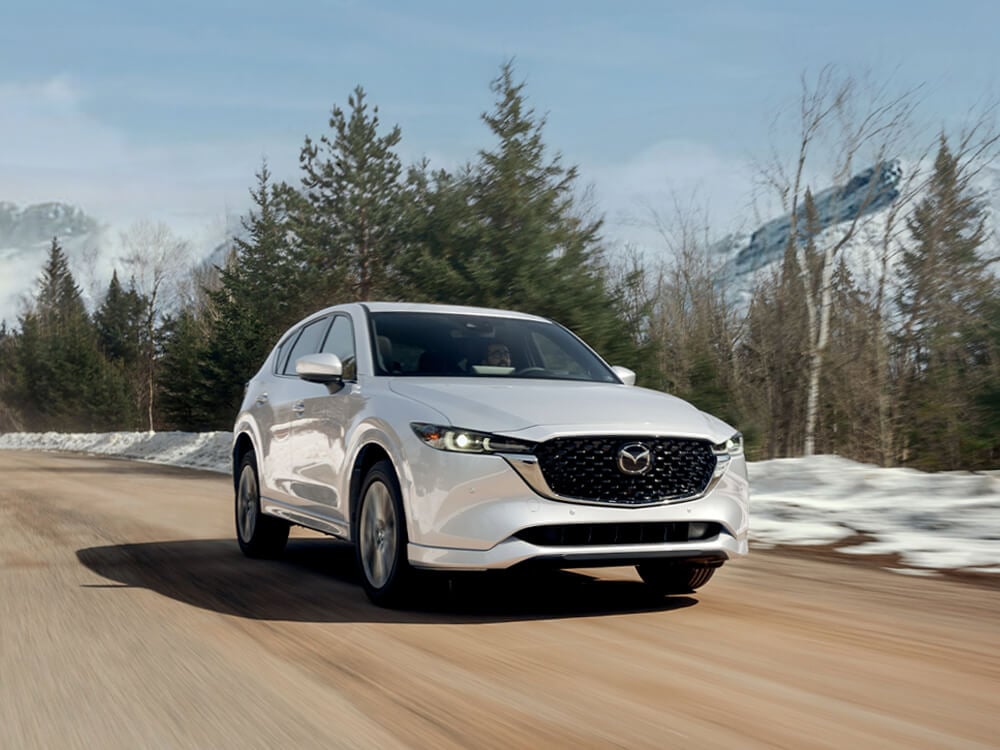 ¾ front Rhodium White Metallic Mazda CX-5 driving on mountain road in winter. 