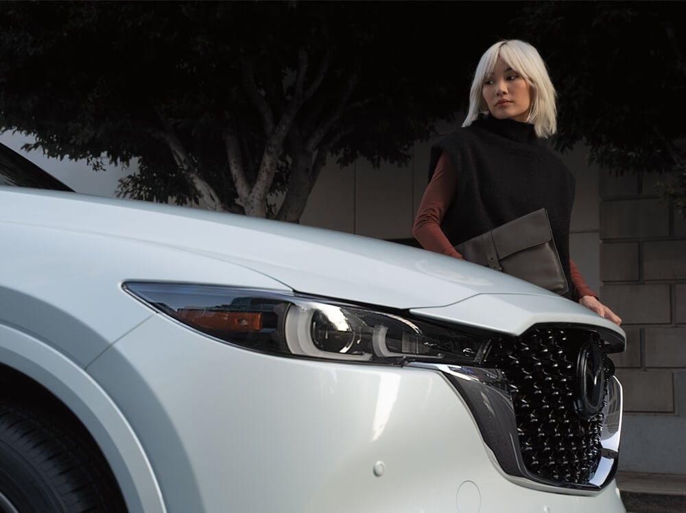 Capot et calandre d’un CX-5 blanc rhodium métallisé au premier plan; une femme élégante regarde en arrière vers le pare-brise.