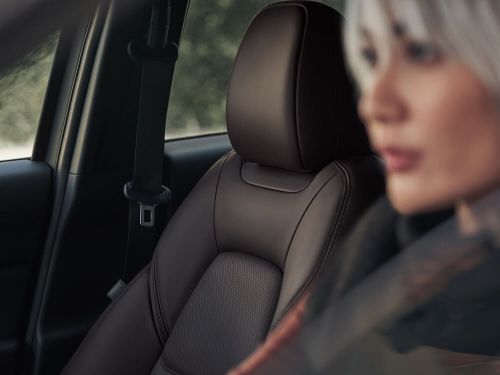 Blurred view of stylish woman driving CX-5 in foreground, Cocoa leather passenger seat details in focus beside her.