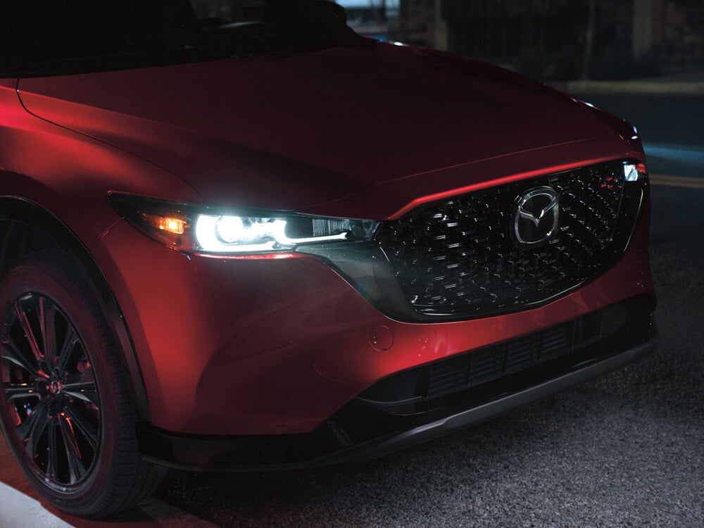 At night, Soul Red Crystal Metallic CX-5 ¾ front right side shows grille, headlight on, 19-inch black metallic alloy wheels. 