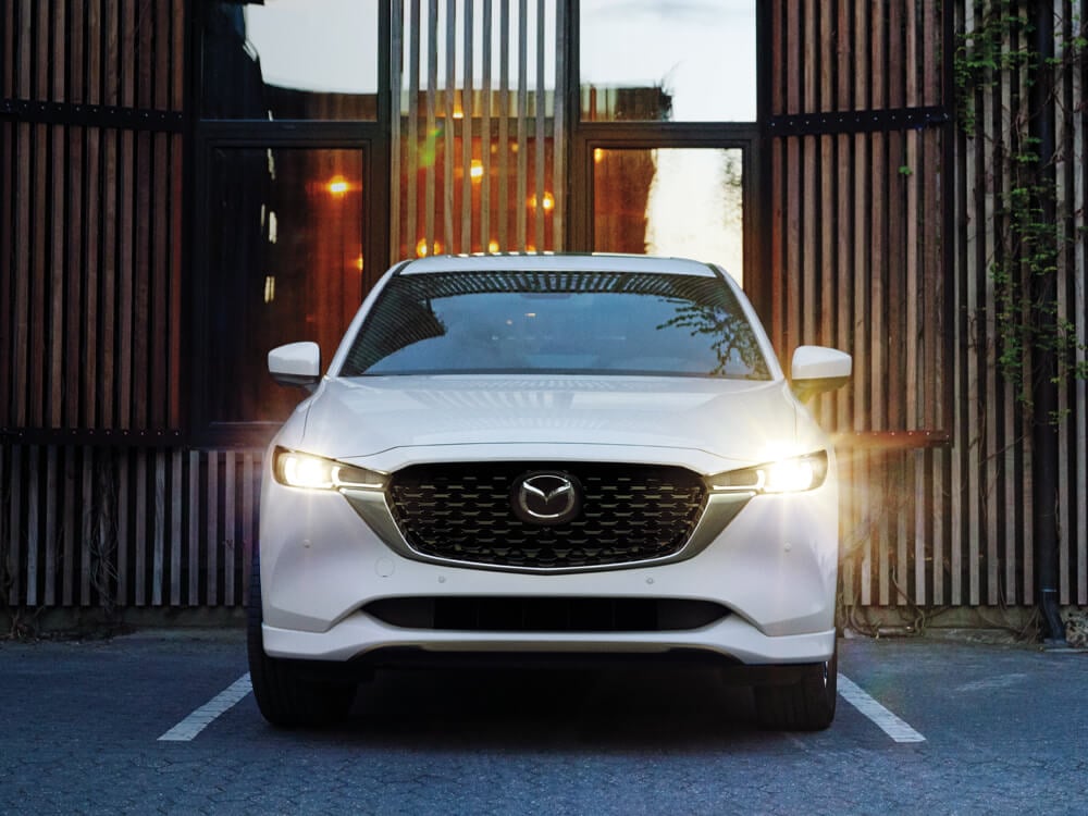 Front on shot of Rhodium White Metallic Mazda CX-5 headlights on, parked in front of stylish minimal building. 