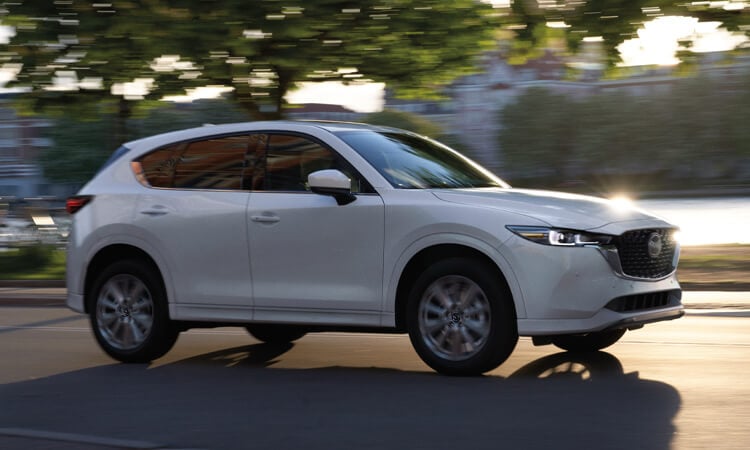 Rhodium white CX-5 driving profile shot, stately building and trees motion blurred in background. 