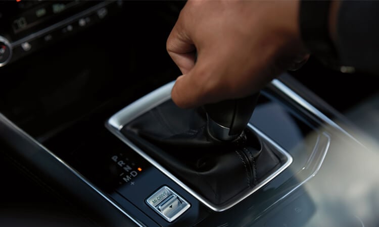 Closeup of CX-5 shifter with driver’s hand on it, and Mi-Drive mode selector switch. 