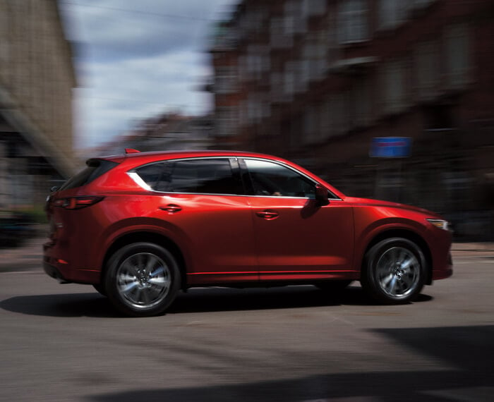 Un CX-5 rouge vibrant cristal métallisé traverse une intersection sur une rue principale par une journée ensoleillée; les bâtiments sont flous sous l’effet du mouvement. 