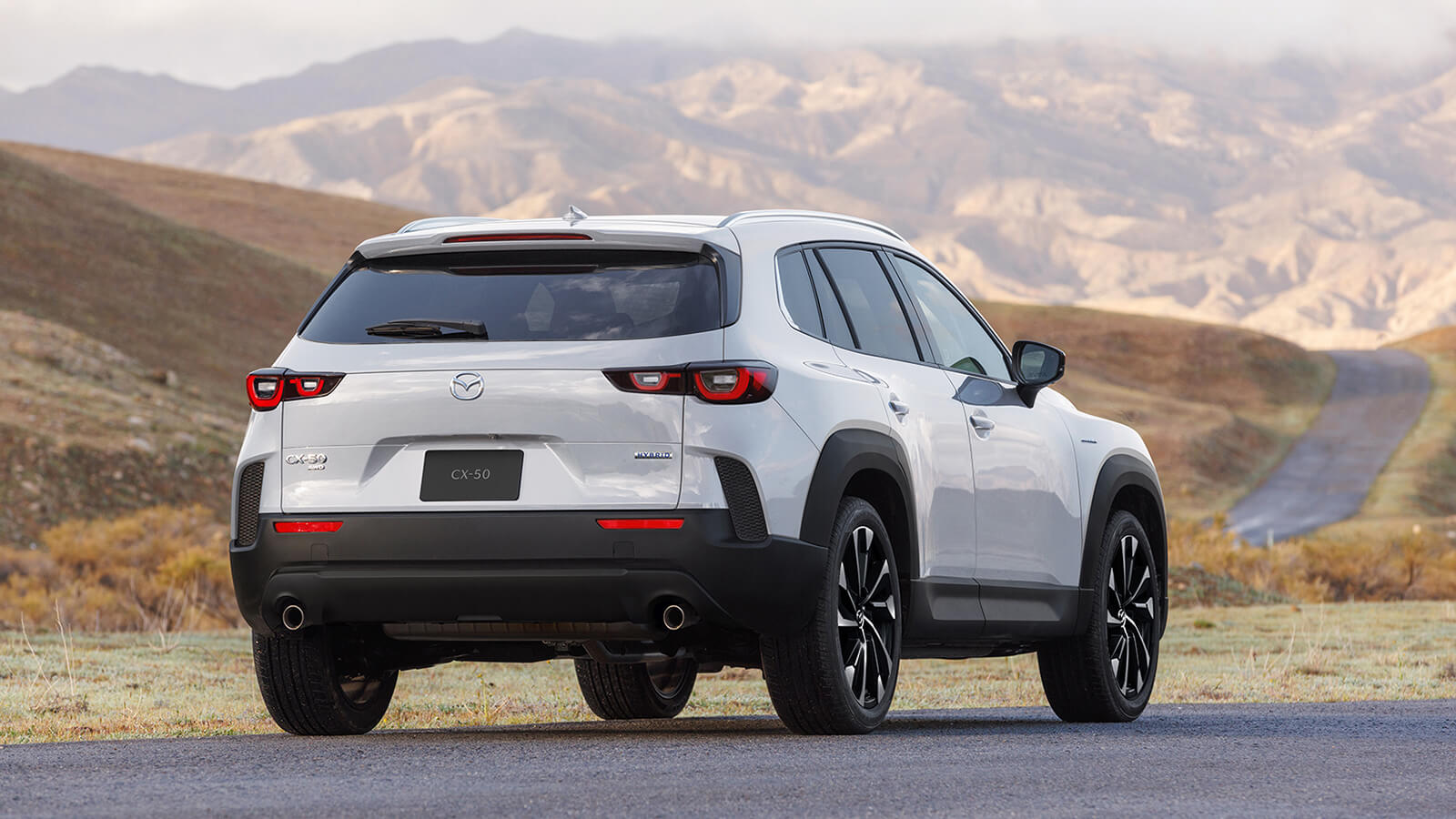 Plan au ¾ arrière d’un CX-50 hybride vent polaire nacré garé sur une route pavée dans un environnement de toundra, avec des montagnes en arrière-plan.