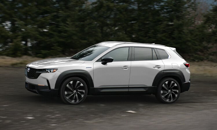 Gros plan de profil d’un Mazda CX-50 hybride vent polaire nacré circulant sur une route avec des arbres en arrière-plan. 