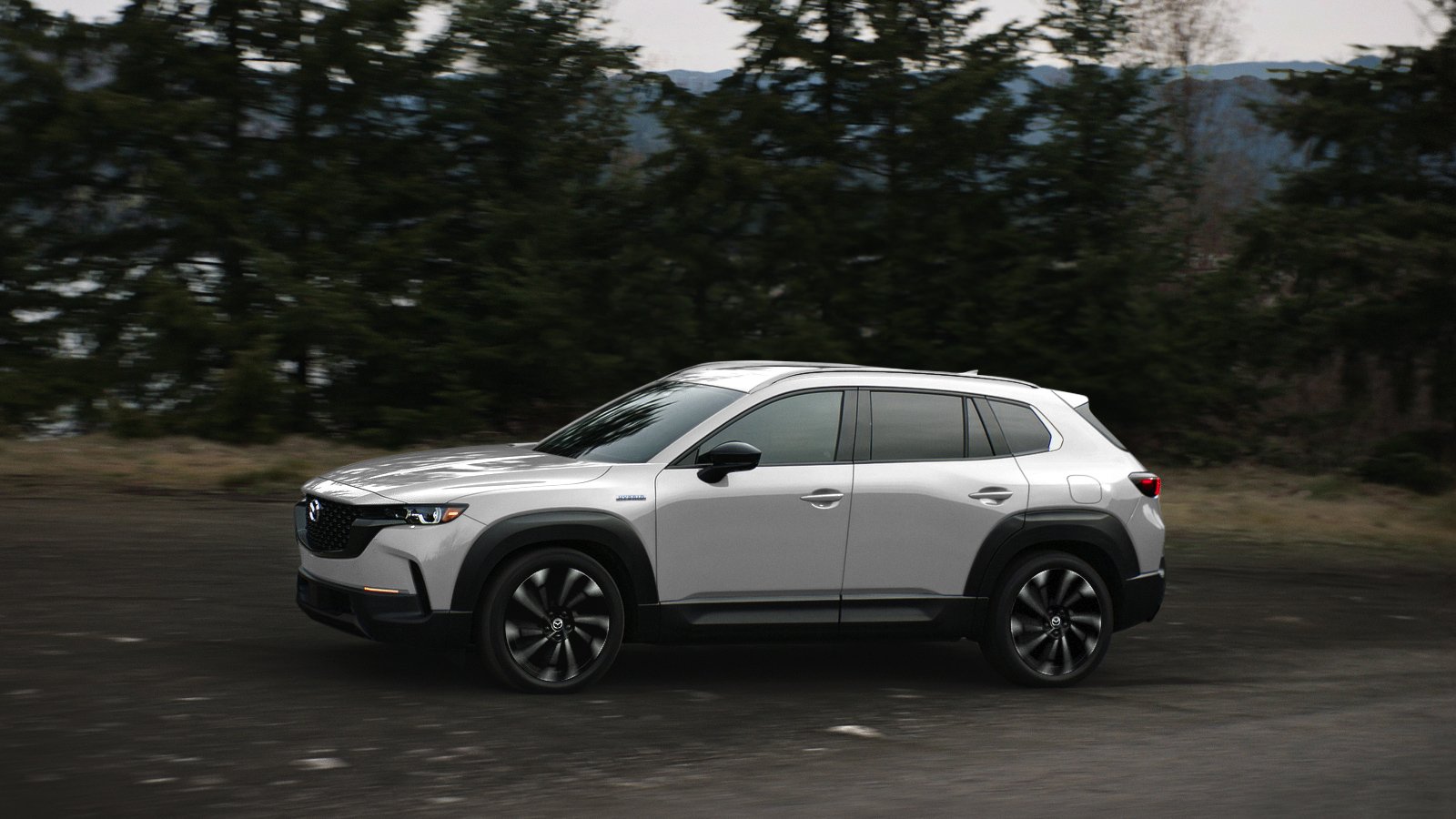 Un CX-50 Vent glacé nacré de profil circulant dans la forêt avec des arbres en arrière-plan; léger flou de mouvement sur les arbres et les jantes du véhicule