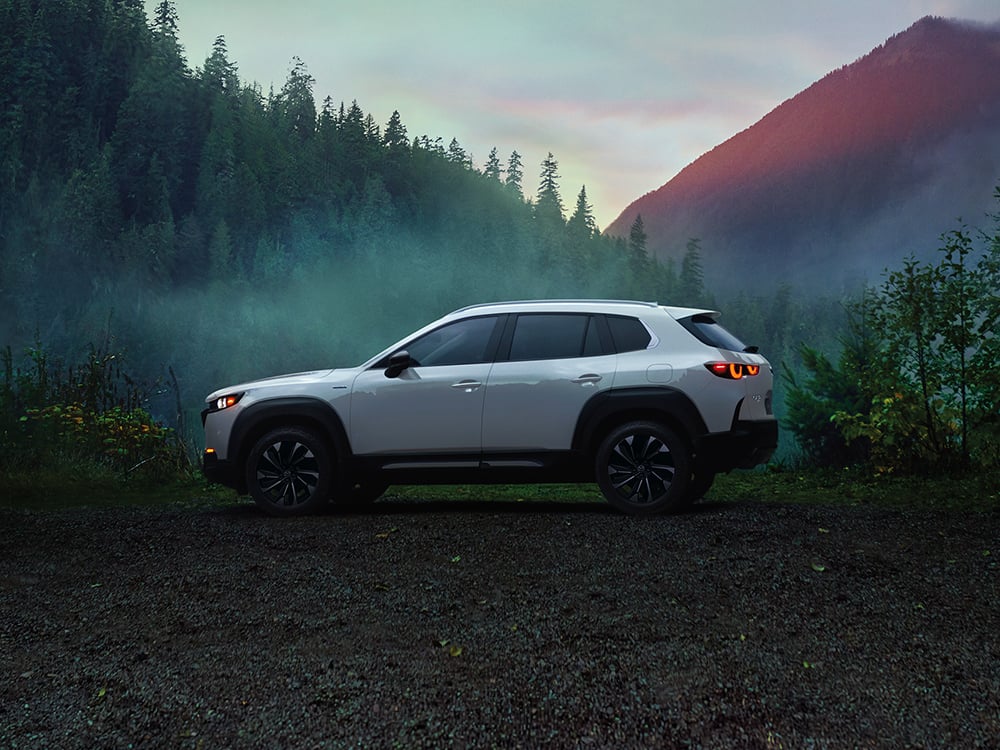 Un CX-50 Vent glacé nacré de profil, garé. On aperçoit une montagne et des arbres en arrière-plan. 