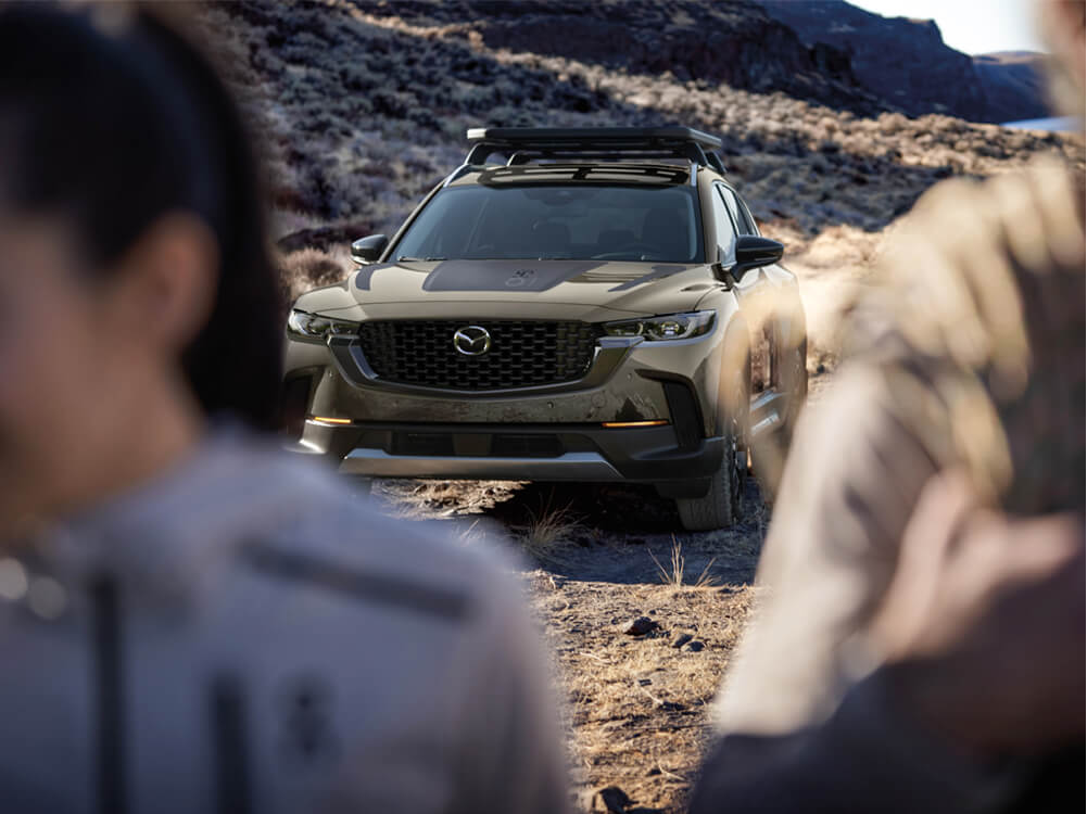 Front view of parked Zircon Sand Metallic Meridian trim CX-50 on gravel path, out of focus people in extreme foreground. 