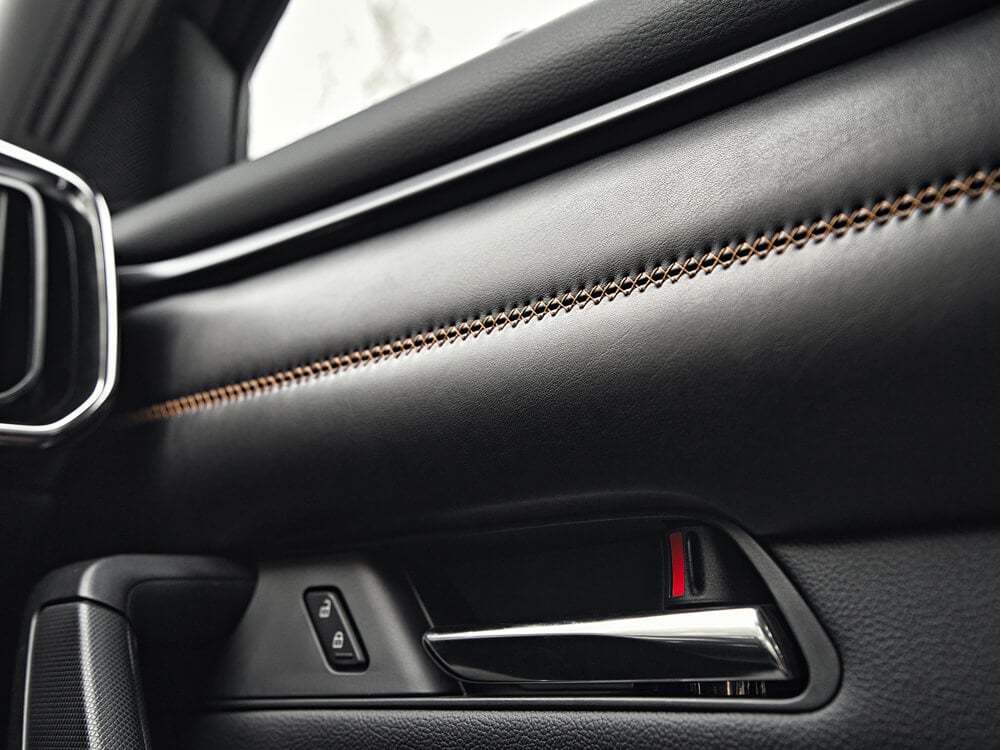 Close up of CX-50 interior front passenger door and handle, black interior with orange stitching details are the focus. 