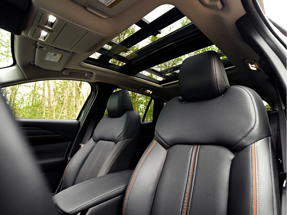 From steering wheel POV, interior shot of two front black leather seats with orange stitching in foreground, looking up through moonroof. 