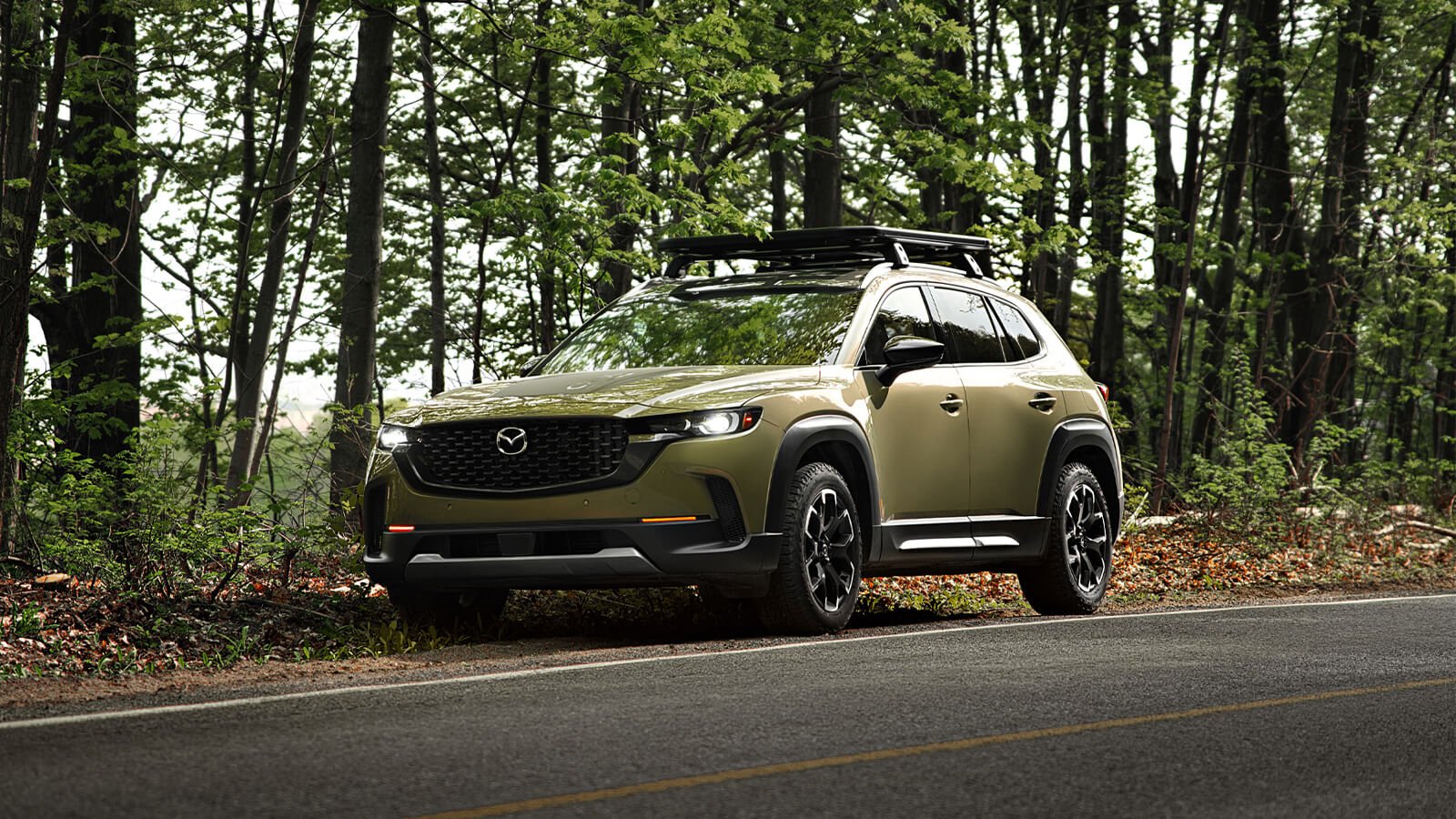 Meridian Trim CX-50 ¾ front, parked on gravel shoulder of paved road in the forest, headlights are on. 