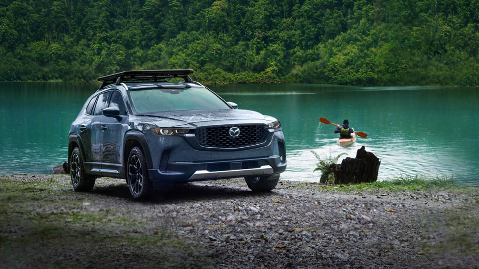 Ingot Blue Metallic CX-50 with roof rack parked ¾ on dirt shore of lake, the driver is in their kayak paddling out into the lake.